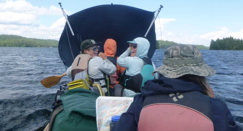 A group of people wearing lifejackets sit in a canoe. They appear to be using some sort of device as a makeshift sail. 